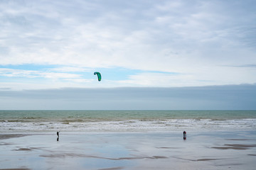 Obraz premium A marée basse, sur une plage de sable, un couple regarde un kitesurfeur. le kitesurf est vert et vole dans un ciel nuageux. La plage reflète les couleurs bleues et grise.