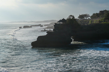 Cliffs in the nearby of Tanah Lot Temple, Bali, Indonesia. There is an arch in the water. The waves are splashing on the cliffs and smaller rocks. Water stays on the flat surfaces. Power of the nature