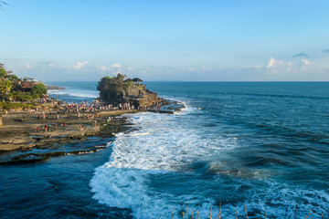 Fototapeta na wymiar Crowds visiting the Tanah Lot Temple, Bali, Indonesia, located on the cliffs by the seashore. The waves are splashing on the cliffs and smaller rocks. Clear and sunny day. Power of the nature