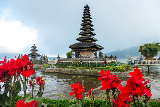 View on the front of main building of Ulun Danu Temple, Bali, Indonesia, through red flowers. Beautiful of Hindu water temple. Sacred place. Discovering new cultures. The temple is surrounded by water