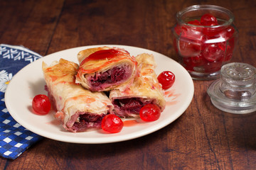 Sour cherry filo pie with syrup poured over it. Candied cherries are placed around pieces of the pie; blue dish towel is lying beside a white plate.