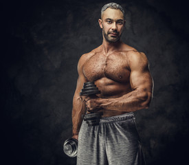 Fototapeta na wymiar Powerful, adult, fit muscular caucasian man coach posing for a photoshoot in a dark studio under the spotlight wearing grey sportswear, showing his muscles and putting up a dumbbells looking confident