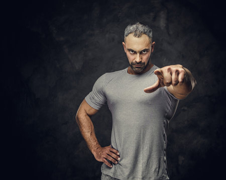 Powerful, adult, fit muscular caucasian man coach posing for a photoshoot in a dark studio under the spotlight wearing sportswear, showing a gesture with his hand forcing to action