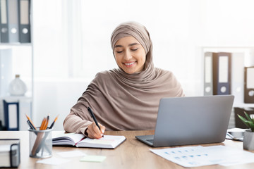 Islamic businesswoman working on laptop in office and writing notes