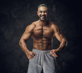 Fototapeta na wymiar Strong, adult, fit muscular caucasian man coach posing for a photoshoot without his shirt in a dark studio under the spotlight wearing sporty shorts, showing his muscles looking powerful and smiling