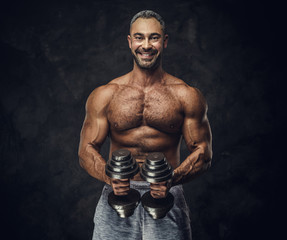 Fototapeta na wymiar Strong, adult, fit muscular caucasian man coach posing for a photoshoot in a dark studio under the spotlight wearing grey sportswear, showing his muscles and putting up a dumbbells looking joyful