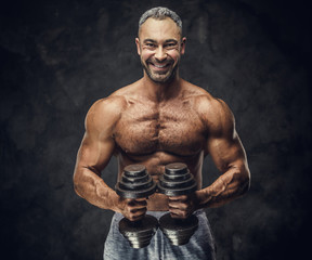 Heavyweight, adult, fit muscular caucasian man coach posing for a photoshoot in a dark studio under the spotlight wearing grey sportswear, showing his muscles and putting up a dumbbells looking
