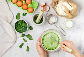 Woman prepares dough for homemade green pancakes for Breakfast. Whisk for whipping in hands. Ingredients on the table - wheat flour, eggs, milk and spinach. Selective focus