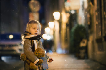 Sweet toddler boy, playing guitar at night in the city with teddy bear toy