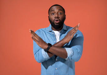 Man raises palms in no or stop gesture. Photo of african man in casual outfit on coral background.