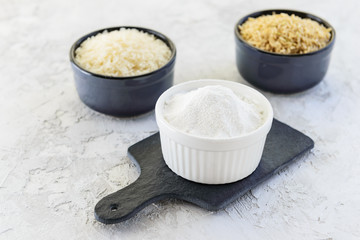 Rice flour in a bowl on a black board on a background of white and brown rice. Gluten free, healthy diet concept.