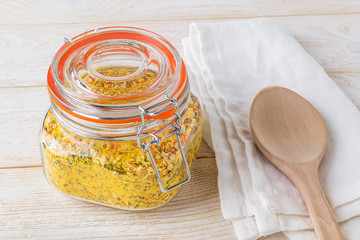 Closed clip top glass jar with universal seasoning and a large wooden spoon on a white wooden kitchen table. Seasoning with salt and dry vegetables. Cook at home.