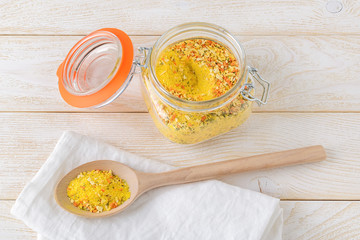 Universal seasoning in a clip top glass jar and in a large wooden spoon on a white wooden kitchen table. Seasoning with salt and dry vegetables. Cook at home.