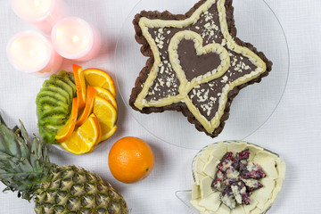 Dessert for a loved one for the holiday. Top view. Star-shaped chocolate cake with a heart inside on white background. Candles, pineapple, slices of orange and kiwi, chopped tofu cheese and chocolate