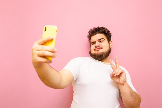 Young man with beard and overweight stands on a pink background, takes a selfie on smartphone camera and shows gesture peace. Funny fat man in white t-shirt makes selfie isolated on pink background