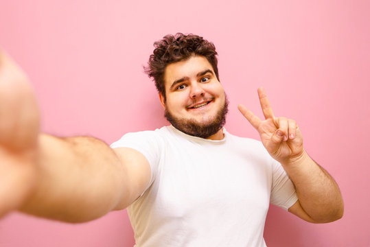 Funny fat man in white t-shirt and beard takes selfies, shows peace gesture and smiles looking into camera, isolated. Cheerful overweight man taking a selfie on a pink background.