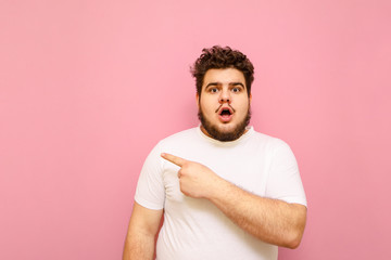Portrait of shocked boy with tire weight on pink background, points his finger away at copy space,...