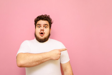 Shocked overweight guy in white t-shirt stands on pink background and points his finger away at copy space. A surprised charismatic big guy looks into the camera and points to a blank space. Isolated