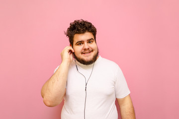 Cheerful charismatic fat man in headphones and white t-shirt isolated. on pink. Happy curly overweight man listening to music in headphones and looking into the camera with a smile on his face.
