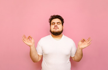 Handsome fat man with curly hair and beard meditates on a pink background with closed eyes wearing a white T-shirt. Authentic overweight guy meditates on a colored background. Isolated