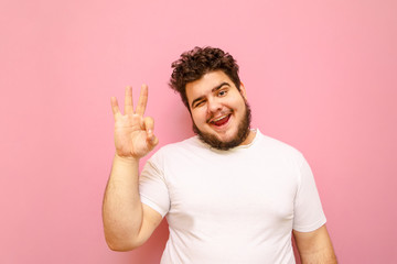 Portrait of a happy guy with curly hair and overweight standing on a pink background, looking into the camera, smiling and showing the gesture Ok, wearing a white T-shirt. Fat man showing "all right"