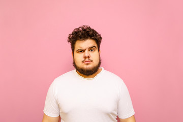 Portrait of a funny fat man in a white t-shirt on a pink background, looking into the camera with a...