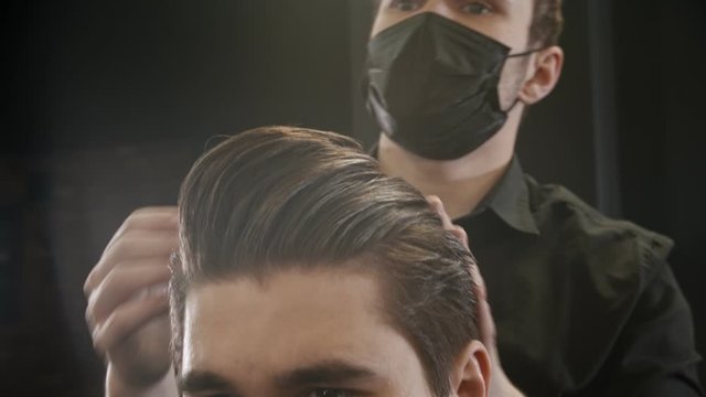 Barber in black mask styling his client hair using a gel