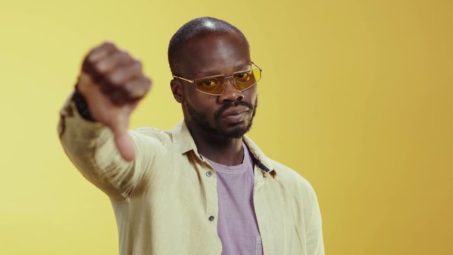 Serious black young millennial man shows refusal gesture with thumbs down disagree negative standing at yellow background. Copy space. Expressions.