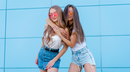 two young hipster woman friends in retro neon sunglasses standing and smiling over blue wall
