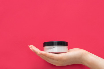 The hand of a girl with a manicure holds a jar of face powder on a pink background. White powder...
