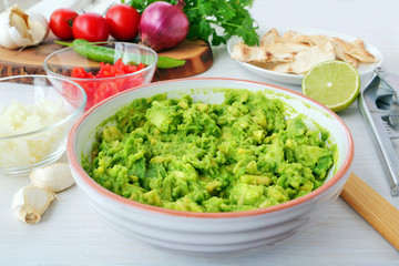 Mashed avocado along with diced onions and tomatoes  in bowls for making avocado dip. Preparing ingredients for guacamole with ingredients and kitchen utensils on table