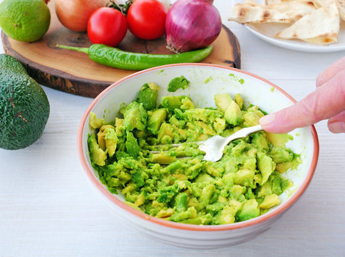 Chef's hand mashes avocado with the back of a fork in a mixing bowl. Making guacamole dip with ingredients on table.