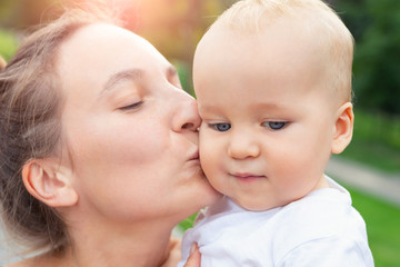 Young adult attractive beautiful caucasian mom kissing little blond toddler boy son during walk in city street park. Mother enjoy happy parenting and childcare . Healthy baby and mama outdoors