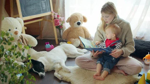 Mom and baby sit on the floor on a soft carpet in the children's room and read a book, dog sleep next to it