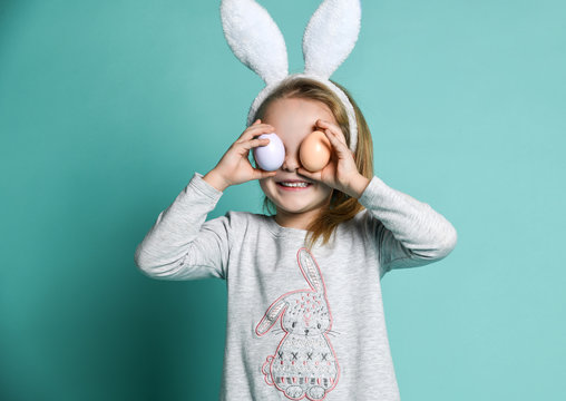 Small happy girl in grey cosy home clothing and decorative fur ears standing and playing with pastel colored eggs