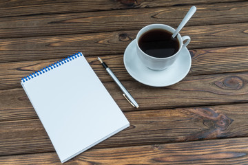 Cup with coffee and a blank notebook with pen on a wooden table. White blank mock up page
