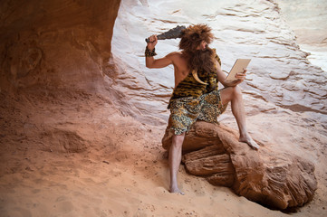 Stone Age luddite caveman scratching his head with a club while looking at his stone tablet outdoors in a weathered rock cave - obrazy, fototapety, plakaty