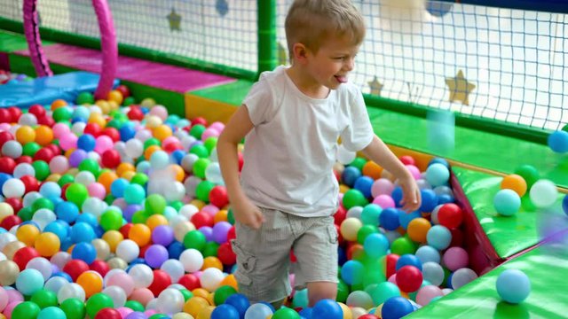 Blonde little boy scatters balls in different sides, lying on multi coloured plastic balls in big dry paddling pool in playing centre. Smiling at camera. Having fun in playroom. Leisure Activity.