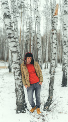 Handsome man standing in forest in winter season. Brutal young male wearing beige jacket over red hoodie and earflap hat posing in snowy forest.