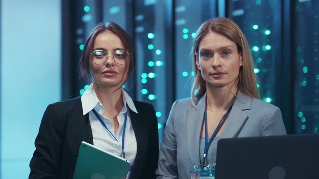 Two women engineers working on laptop inside secure server room. Team portrait of confident female technicians collaborating in futuristic network data center.