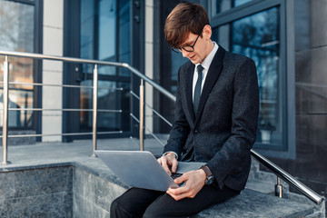 Confident young businessman, uses a laptop for work, sitting on the street against the background of a glass business center