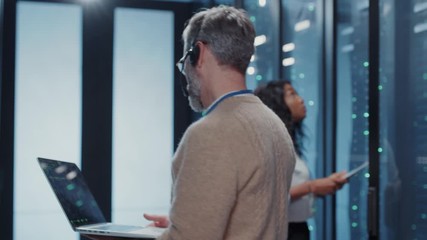 Caucasian IT operator looking at server racks collecting important data on laptop. His pretty female colleague doing data transfer operations for computer security. Teamwork. - Powered by Adobe