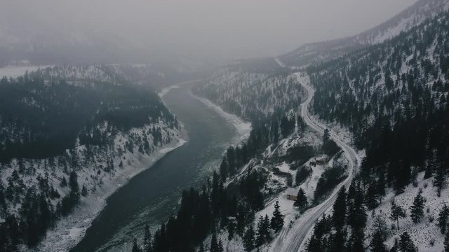 Aerial view of the winter river, falling down snow and the mountains covered with the pine trees