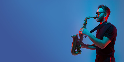 Young caucasian jazz musician playing the saxophone on gradient blue-purple studio background in...