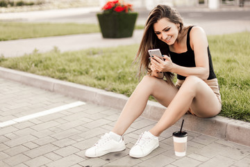 Beautiful young woman using phone and drink coffee sitting on the curb in the street.