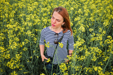Naklejka na ściany i meble Female photographer wearing a shirt with stripes scouts around in the middle of a rapeseed field.