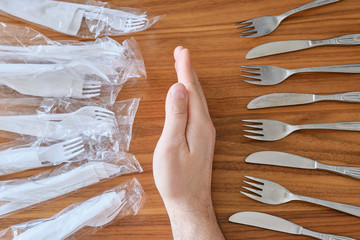 Plastic versus reusable cutlery concept: Man hand between disposable plastic sets and a stainless steel fork / knife set, on a walnut table top.
