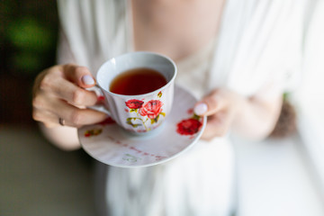 Close-up of a mug with a drawing on a saucer in the hands of a young woman. The smell of tea in a vintage mug with coffee in the hands of a girl in Bathrobe on the background of an expensive interior