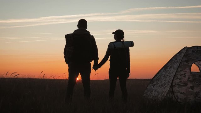 Silhouette tourist couple standing holding hands and watching the sunset near tent in vacation. Camp rest lifestyle. Tourism and camping concept.