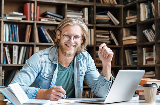 Portrait Young Bearded Male College Student Glasses Sit At Home Library Desk Handwrite Notebook Hold Pen Make Notes From Textbook Laptop Prepare Project Study Online Exam Test Web E Education Concept.